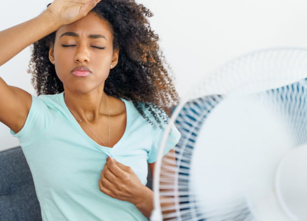 A woman in a blue shirt holds her hand to her head as she sits in front a fan to cool off